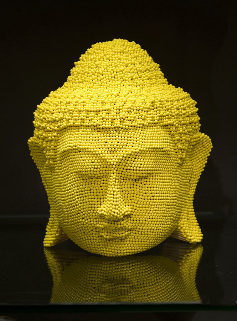 Yellow sculpture of a Buddha head made entirely out of match sticks