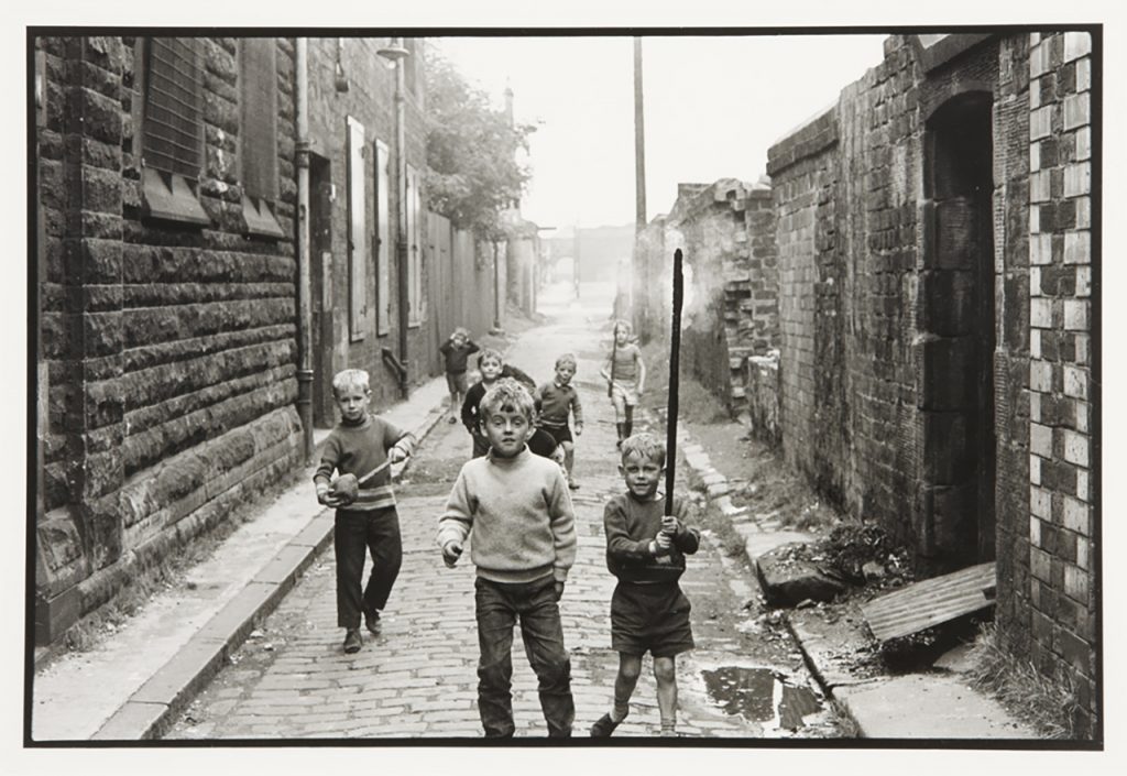 Black and white photograph of children in the street starting directly at the camera. Some of the children are holding sticks