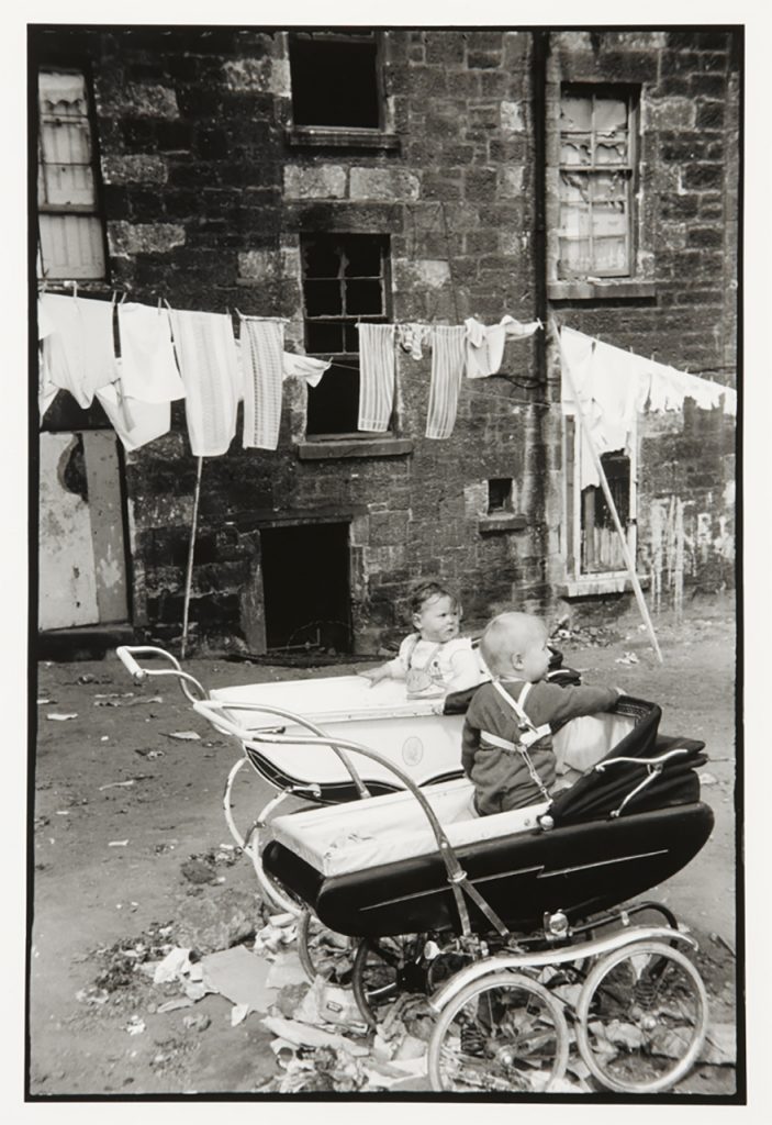 Black and white photograph of two prams with babies in parked outside. In the background are buildings and a line of laundry hung out to dry.