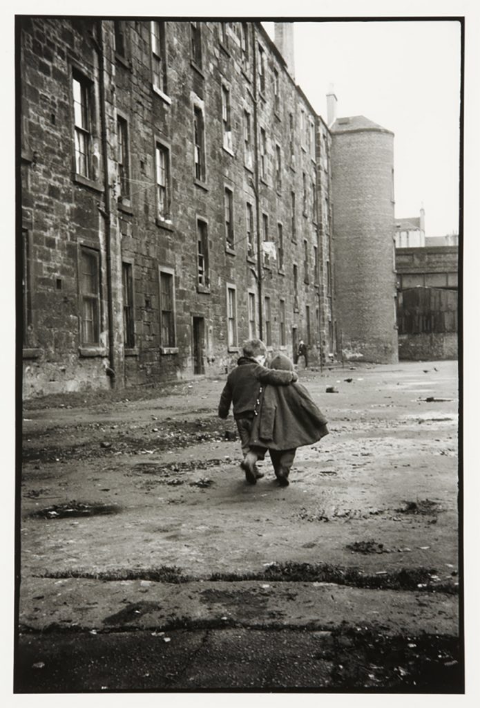 Black and white photograph of two children walking arm in arm past some large tenement buildings.