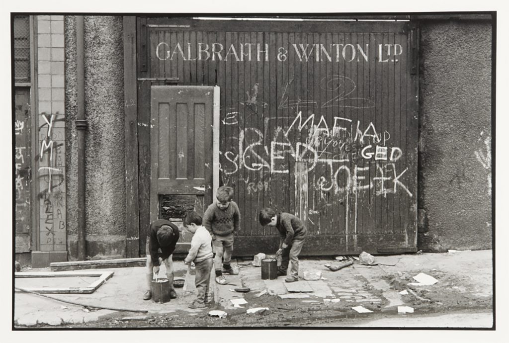 Black and white photograph of some children painting a wall covered in graffiti.