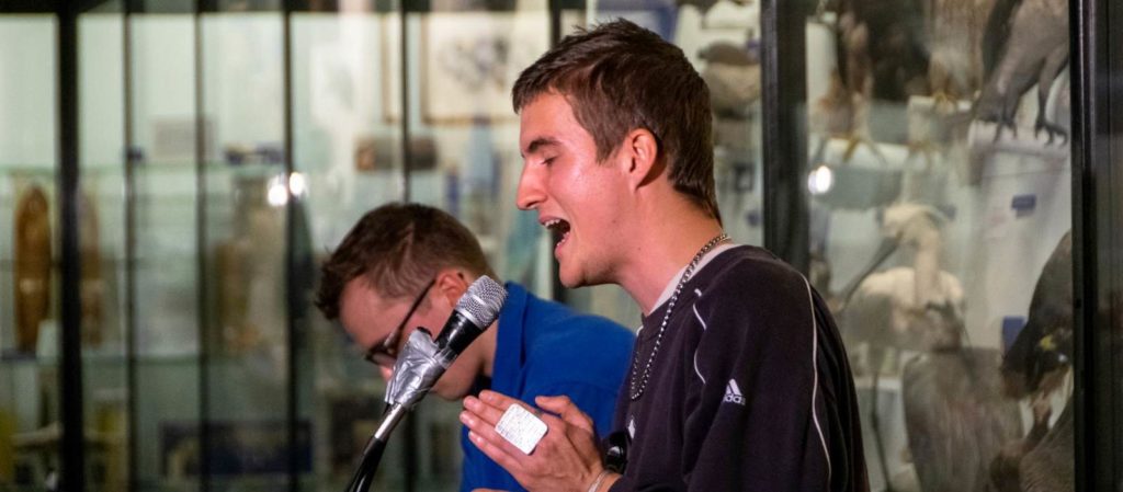 two male students in centre of image, one singing into a microphone in front with hands clasped together and one with his head dipped behind. In the surrounding background can be seen cases of animal specimens in the Bell Pettigrew Museum of natural history.