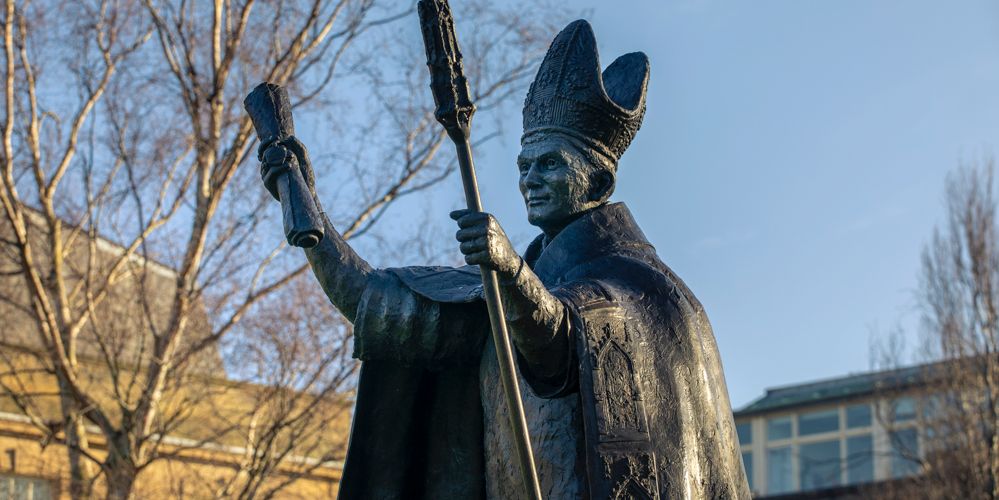 metal statue of Bishop Henry Wardlaw in centre of photo and in focus, in Bishop's clothing holding document in right hand and mace in the left. Bare tree to left of image and buildings to left and right in background. 