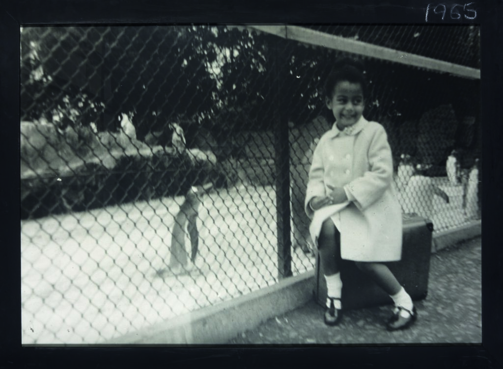 Black and white photograph of a young child smiling whilst sat in front of an enclosure of penguins.