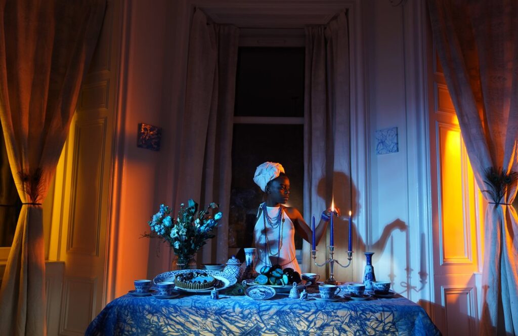 A women stands by a dining table lighting a candle. Chinaware, flowers, and blue cloth cover the table as orange lights come through the windows from the left and right. 